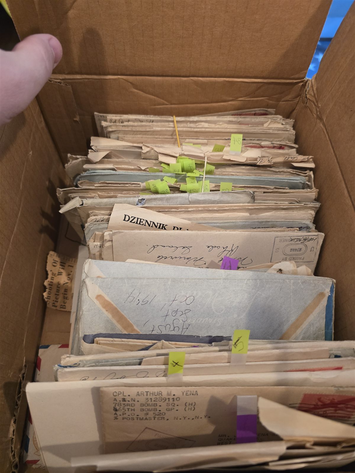 Looking down into the box: tightly packed rows of 1940s envelopes flagged with green and purple archival tabs, a Polish-language newspaper folded among them.